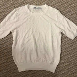Vit stickad tröja från Zara - En stilren vit stickad tröja från Zara med rund halsringning och korta ärmar. Perfekt för dig som gillar en enkel och clean look. Tröjan har ribbade muddar vid ärmslut och nederkant för en snygg passform.