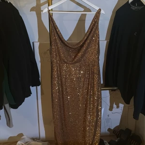Glittrig guldklänning från Undress - Supersnygg långklänning från Undress i guld med glittriga paljetter. Klänningen har en draperad urringning, smala axelband och öppen rygg. Perfekt för dig som vill sticka ut och glänsa på festen. Materialet är mjukt och följsamt med lyxig känsla. Kommer även med matchande midje band.