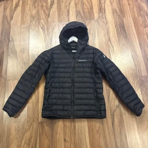 Svart dunjacka Peak Performance L - Svart dunjacka från Peak Performance med huva och dragkedja framtill. Jackan är quiltad med horisontella sömmar och har två fickor med dragkedja. Perfekt för kyliga dagar och har en stilren look med diskret logga på bröstet.