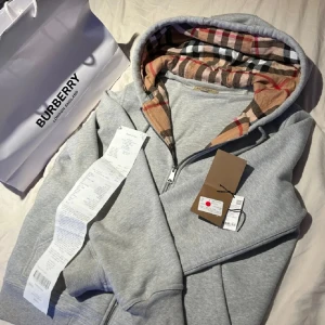 Grå Burberry hoodie med rutigt foder - Säljer en grå hoodie från Burberry med dragkedja och klassiskt rutigt foder i huvan. Tröjan har långa ärmar, fickor framtill och ikoniskt Burberry-mönster på insidan av huvan. Perfekt för dig som gillar stilrena och lyxiga streetwear-plagg.