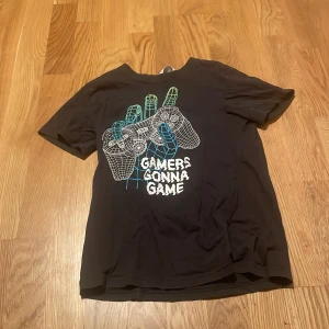 Svart gamer t-shirt från H&M - Svart t-shirt från H&M med coolt tryck av en hand som håller en spelkontroll och texten 'Gamers Gonna Game' på framsidan. Klassisk passform med rund hals och korta ärmar. Perfekt för dig som älskar gaming och vill ha en chill stil.