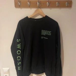 Svart Nike sweatshirt med grön text - Svart sweatshirt från Nike med grön text på bröstet och stort 'SWOOSH' tryckt längs ena ärmen. Klassisk crewneck-modell med lång ärm och relaxed fit. Perfekt för dig som gillar sportig och clean streetstyle.