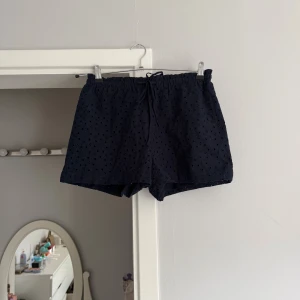Mörkblå broderade shorts från H&M - Säljer ett par mörkblå shorts från H&M i bomull med broderade blommor och hålmönster. De har elastisk midja med snörning och en loose fit som är perfekt för varma dagar. Supersöta och lätta att matcha med allt.