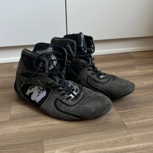 Gorilla Wear svarta träningsskor - Säljer ett par svarta och grå träningsskor från Gorilla Wear med högt skaft och snörning. Skorna har en cool gorilla-logga på sidan och är gjorda i mocka och syntetmaterial. Perfekta för gymmet eller styrketräning.
