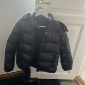 Svart pufferjacka från Polo Ralph Lauren - Mycket fin jacka, bra skick Svart pufferjacka från Polo Ralph Lauren med klassisk logga på bröstet och stor etikett på sidan. Jackan har dragkedja framtill och hög krage. Perfekt för kalla dagar och riktigt snygg till streetwear-stilen.