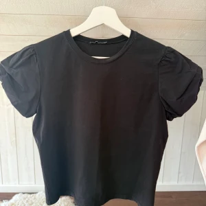 Svart puffärm topp från Zara - Säljer en svart topp från Zara med puffade korta ärmar och rund halsringning. Toppen är gjord i mjuk bomull och har en clean, stilren look som är enkel att matcha. Perfekt för dig som gillar minimalistisk stil med en twist på ärmarna.