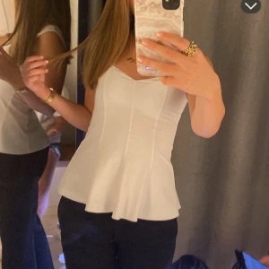 Vit bandeau med breda axelband - Säljer en vit bandaeu-topp med breda axelband och markerad midja. Toppen har en lätt utsvängd peplum-nederdel och en snygg v-ringning framtill. Materialet är slätt och känns bekvämt mot huden. Perfekt att styla med jeans eller kjol för en trendig look.