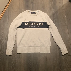  stickad tröja Morris  - Ljusgrå stickad tröja från Morris med mörkblå panel och vit text framtill. Tröjan har rund hals, ribbade muddar och är långärmad. Perfekt för dig som gillar klassisk och sportig stil.