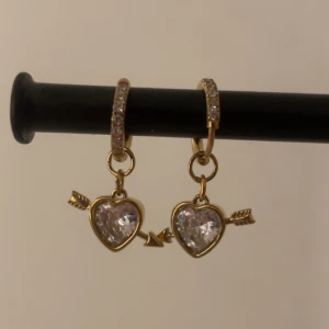 Guldiga örhängen med hjärtan och pil vit - Superfina örhängen i guldton med gnistrande stenar på ringen och hängande hjärtformade berlocker med pil igenom. Hjärtana är dekorerade med klara stenar som ger extra bling. Perfekta för dig som gillar söta detaljer och vill sticka ut.