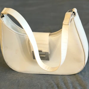 Vit axelväska med silverdetalj - Stilren vit axelväska i skinnimitation med snyggt silverfärgat spänne framtill. Väskan har en klassisk, rundad form och justerbar axelrem. Perfekt storlek för det viktigaste och enkel att matcha med olika outfits.