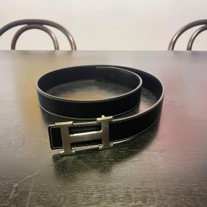 Svart skärp med H-spänne - Svart skärp  med ikoniskt H-spänne i silverfärgad metall. Bältet är tillverkat i exklusivt skinn och har vita kontrastsömmar längs kanterna. Perfekt accessoar för att lyfta din outfit med en lyxig touch.