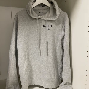 Grå hoodie från A.P.C.  - Säljer nu en skitfet ljusgrå hoodie från A.P.C. med svart tryck på bröstet. Klassisk modell med huva och dragsnören, samt långa ärmar. Tillverkad i mjuk bomull för en skön och avslappnad stil. Storlek L men passar M eller till och med S eftersom den är mindre i storleken. Skriv om minsta lilla fundering!