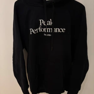 Svart hoodie från Peak Performance - Svart hoodie från Peak Performance med vit broderad logga på bröstet och klassisk känguruficka. Tröjan har huva med snörning och är tillverkad i mjuk bomullsblandning. Perfekt för chill dagar eller när du vill ha en clean streetstyle-look.