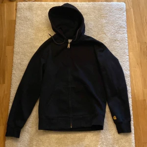 Svart hoodie från Carhartt med dragkedja - Svart hoodie från Carhartt med gul broderad logga på ärmen. Tröjan har huva med snörning, hel dragkedja framtill och känguruficka. Perfekt för dig som gillar streetwear och enkel stil.