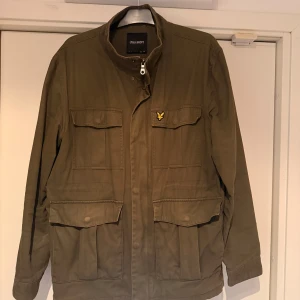 Olivgrön parkajacka från Lyle & Scott - Snygg olivgrön parkajacka från Lyle & Scott med klassisk gul logga på bröstet. Jackan har fyra stora fickor framtill, ståkrage och dragkedja med knappstängning. Tillverkad i bomullsmaterial och har långa ärmar med knappdetalj vid mudden.