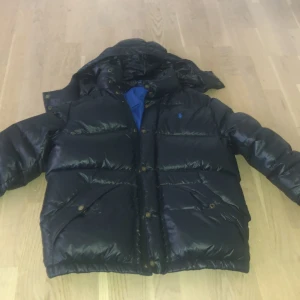 Svart dunjacka från Polo Ralph Lauren - Säljer en svart dunjacka från Polo Ralph Lauren med blått foder och huva. Jackan har puffad design, tryckknappar och dragkedja framtill samt två fickor med knappstängning. Perfekt för kalla dagar och har en klassisk look med diskret logga på bröstet.