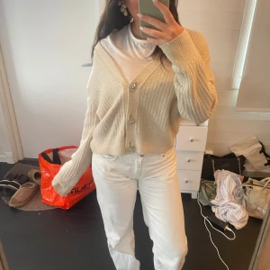 Beige ribbad kofta med pärlknappar - Superfin beige ribbstickad kofta från H&M med v-ringning och två glittriga pärlknappar framtill. Modellen är croppad och har långa ärmar. Perfekt att slänga över en t-shirt för en chill look.