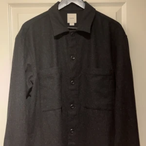 Svart overshirt från Calvin Klein L - Stilren svart overshirt från Calvin Klein i storlek L. Jackan har klassisk krage, raka linjer, två stora bröstfickor och knäppning framtill. Perfekt för lager-på-lager och enkel att matcha med allt. Materialet känns mjukt och har en ullig look.