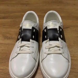 Valentino Garavani Rockstud sneakers - Snygga Valentino Garavani Rockstud sneakers i vitt skinn med svart detalj över snörningen och ikoniska nitar på hälen. Klassisk rund tå, platt sula och vita skosnören. Perfekta för dig som vill ha en stilren men edgy look.