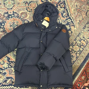 Mörkblå dunjacka från GANT med huva - Säljer en mörkblå dunjacka från GANT med stor, mysig huva och snörning. Jackan har quiltad design, dragkedja och tryckknappar framtill samt ett brunt GANT-märke på ärmen. Perfekt för kalla dagar och riktigt snygg till streetwear.