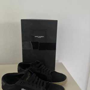Svarta Saint Laurent sneakers - Snygga svarta sneakers från Saint Laurent Paris med klassisk låg profil och vit sula. Skorna har snörning, rund tå och diskret logga på sidan samt plös. Tillverkade i mocka och skinn för en lyxig känsla. Levereras med originalkartong och dustbag.