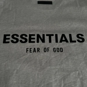 Grå Essentials Fear of God t-shirt oversized  - Snygg grå t-shirt från Fear of God Essentials med svart tryck både fram och bak. Klassisk rund hals och korta ärmar. Materialet är mjuk bomull som känns skönt mot huden. Perfekt för dig som gillar stilrena streetwear-plagg med tydlig logga.