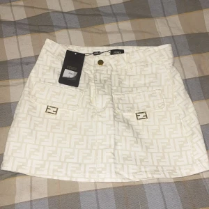 Beige kortkjol med fendi prints - Snygg beige kortkjol från Fendi med ikoniskt FF-mönster i beige. Kjolen har framfickor med guldfärgade Fendi-loggor, knappstängning och bälteshällor. Materialet är i jeans, kjolen passar inte mig som har storlek L men passar bra på någon som är S-M