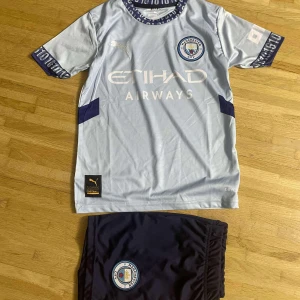 Manchester City De Bruyne tröja Puma - Säljer en ljusblå Manchester City fotbollströja med De Bruyne 17 på ryggen, tillverkad av Puma. Tröjan har korta ärmar med mörkblå detaljer och klubbmärke på bröstet. Materialet är lätt och andas, perfekt för träning eller match. Matchande mörkblå shorts med klubbmärke ingår.