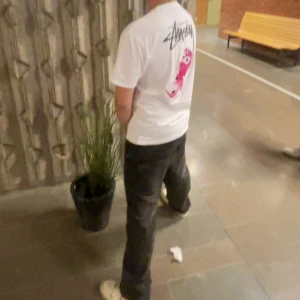 Vit Stüssy t-shirt med tryck - Vit t-shirt från Stüssy med svart logga på bröstet och stort tryck med rosa panter på ryggen. Klassisk passform och rund hals. Perfekt för dig som gillar streetwear och vill sticka ut med en ikonisk design.