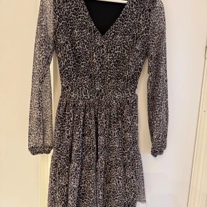 Leopardmönstrad klänning med v-ringning - Snygg kort klänning med leopardmönster och v-ringad hals. Klänningen har långa ärmar i transparent tyg och markerad midja för en feminin siluett. 