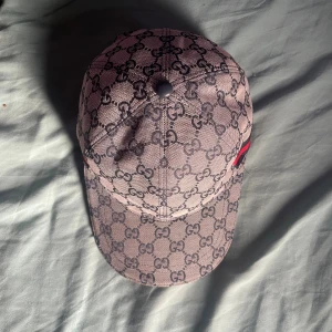 Gucci keps med GG-mönster och röd detalj - Säljer en snygg keps från Gucci i grått med klassiskt GG-monogram över hela kepsen. Kepsen har en röd och marinblå rand på sidan och justerbar rem bak. Tillverkad i bomull och polyester, perfekt för dig som vill sticka ut med en lyxig accessoar.