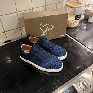 Christian Louboutin skor  - Snygga mörkblå sneakers från Christian Louboutin i mocka med coola nitar på tån och klassisk röd sula. Skorna har snörning och vit platt sula, perfekt för dig som vill sticka ut med exklusiv streetstyle.
