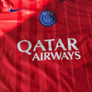 PSG röd matchtröja Nike L - Paris Saint-Germain röd fotbollströja från Nike med blå detaljer och vita tryck. Tröjan har vertikala ränder, PSG-emblem, Qatar Airways sponsortryck och nummer 87 Joao Neves på ryggen. Materialet är lätt och ventilerande polyester, perfekt för träning eller match.