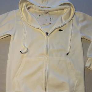Vit  Lacoste hoodie  - Vit hoodie  Lacoste med klassisk krokodil-logga på bröstet. Tröjan har huva med snörning, dragkedja framtill och känguruficka. Tillverkad i mjuk bomull och har ribbade muddar vid ärmslut och nederkant. Perfekt för en clean och sportig look.