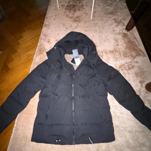 Svart dunjacka från Lexington  - Svart dunjacka från Lexington med huva, dragkedja och snörning nedtill. Jackan har två fickor med dragkedja och beige foder. Perfekt för kalla dagar och har en clean, modern look som funkar till allt. En varm jacka som kommer vara perfekt nu till vintern. Jackan är inte använd och har lapparna kvar, men ändå har den en liten defekt som man kan se på sista bilden. Men de går säker att fixa enkelt. Nypris= runt 5000kr. Mitt pris 2499kr. Kommer inte sänka priset då de är riktigt bra pris