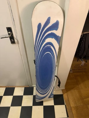 Vortex snowboard med blått mönster - Snowboard från Vortex med vitt och blått grafiskt mönster och två blå ränder på ovansidan. Brädan har svarta och gråa bindningar och en clean, sportig look. Perfekt för dig som vill sticka ut i backen med stilrena detaljer. Lite slitage på nosen men annars bra skick. Perfekt för nybörjare!!
