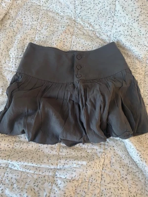 Grå kortkjol från Diesel med knappar - Supersnygg grå kortkjol från Diesel med bred midja och tre dekorativa knappar framtill. Kjolen har en ballongliknande form och är tillverkad i mjukt, lätt material som ger ett coolt fall. 