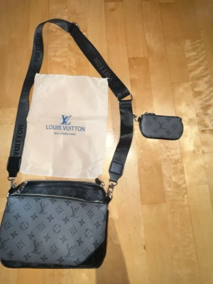 Axel väska - Snygg Louis Vuitton axelväska i elegant grå/svart monogramdesign. Levereras med en justerbar Louis Vuitton axelrem och en matchande miniväska. Ett modernt och mångsidigt plagg, perfekt för vardagsbruk eller speciella tillfällen. Väskan är i gott skick och har tagits väl om hand. Tidlös design som matchar alla outfits och ger en lyxig touch. Tveka inte att kontakta mig om du har några frågor eller vill ha fler bilder.