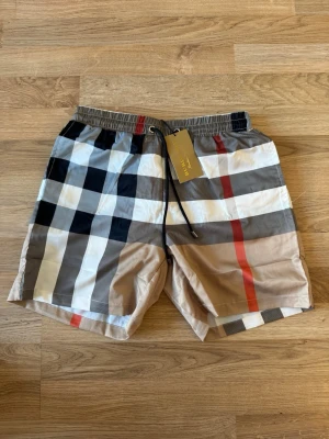 Rutig Burberry shorts med snörning - Säljer ett par klassiska Burberry shorts med ikoniskt rutmönster i beige, svart, vit och röd. Shortsen har elastisk midja med snörning och en bakficka. Perfekta för sommaren och riktigt snygga till streetwear-looken. STORLEK S