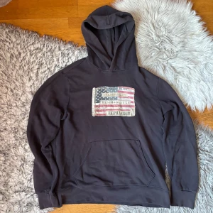 Mörkblå denim supply Ralph lauren tröja med tryck - Ralph Lauren Denim & Supply hoodie  Snygg och bekväm hoodie från Ralph Lauren Denim & Supply. Mörk färg med klassiskt amerikanskt flaggtryck på bröstet. Tröjan har huva och magficka framtill. Perfekt för vardagsbruk och en avslappnad stil.  Skick: Bra begagnat skick, normalt slitage Passform: Klassisk hoodie-passform Färg: Mörk (svart/mörkgrå)  