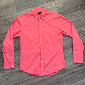 Rosa skjorta från Polo Ralph Lauren - Säljer en snygg rosa skjorta från Polo Ralph Lauren i slim fit. Skjortan har klassisk button-down krage, vita knappar framtill och ett blått broderat logomärke på bröstet. Tillverkad i mjuk bomull och har långa ärmar med knapp vid manschetten.