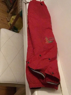 Röda byxor från Cross Active - Röda byxor från Cross Active med broderad logga och detaljer i gult och grått. Byxorna har tryckknappar och resår i benslut, samt dekorativa sömmar. Perfekta för dig som gillar sportig och färgstark stil.