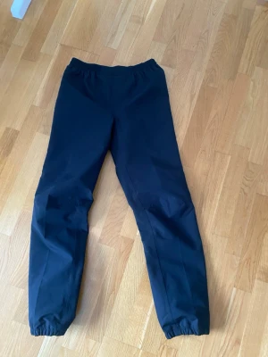 Svarta täckbyxor med resår - Säljer ett par svarta täckbyxor med resår i midjan och vid bensluten. Byxorna har en enkel och clean look, med en reflexdetalj på ena benet för extra synlighet. Perfekta för kalla dagar och utomhusaktiviteter.