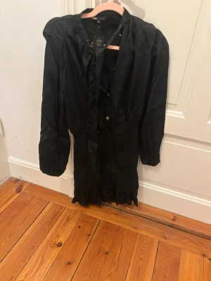 Svart kortklänning från Maje - Supersnygg svart kortklänning från Maje med långärmade puffärmar, knytband i halsen och smockad midja. Kjolen har volangkant nertill och klänningen är gjord i ett glansigt material. Perfekt för dig som vill ha en chic och trendig look.