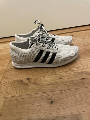 Vita Adidas Los Angeles sneakers - Säljer ett par vita Adidas Los Angeles sneakers med svarta ränder och mesh-överdel. Skorna har snörning och en lätt, ventilerande design som passar perfekt till streetwear. Klassisk Adidas-look med logga på plösen och sidan.