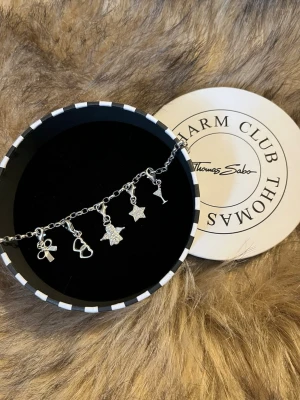 Snygg silverfärgad charm-armband Thomas Sabo - Charm-armband från Thomas Sabo i silverfärg med fem olika hängen: rosett, hjärtan, ängel, stjärna och cocktailglas. Kedjan är grov och armbandet har en rund bricka vid låset. Perfekt accessoar för att lyfta din stil. Värde 1044kr. 18cm 