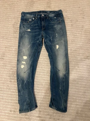 Blå jeans från G-Star RAW - Säljer ett par blå jeans från G-Star RAW med slitna detaljer och distressed look. Jeansen har klassisk femficksmodell, raka ben och tydliga sömmar. Materialet är robust denim i bomull och passformen är normal. Perfekta för en avslappnad stil.