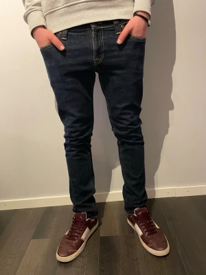 Nudie Jeans Tight Long John blå jeans - Säljer ett par mörkblå Nudie Jeans Tight Long John med klassisk femficksdesign och snygga kontrastsömmar på bakfickorna. Jeansen är tillverkade i 98% ekologisk bomull och 2% elastan för extra stretch och bekväm passform. Lägg gärna prisförslag!!