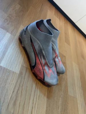 Nike fotbollsskor grå och röd - Säljer ett par gråa Nike fotbollsskor med röda och svarta detaljer. Skorna har en strumpliknande passform utan snörning och en modern design med mönstrade sidor. Perfekta för dig som vill ha stil på planen. Skorna går att få renare men har inte haft ork för det.