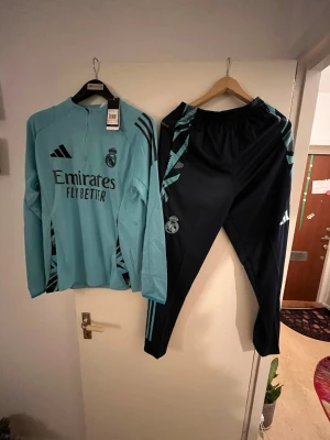 Real Madrid träningsset Adidas - Säljer ett Real Madrid träningsset från Adidas med långärmad turkos tröja och svarta byxor med turkosa detaljer. Setet har klubbmärke och Adidas-logga, samt snygga ränder längs ärmar och ben. Perfekt för dig som älskar fotboll och vill träna i stil. Nytt skick finns i storlekarna S-M-L